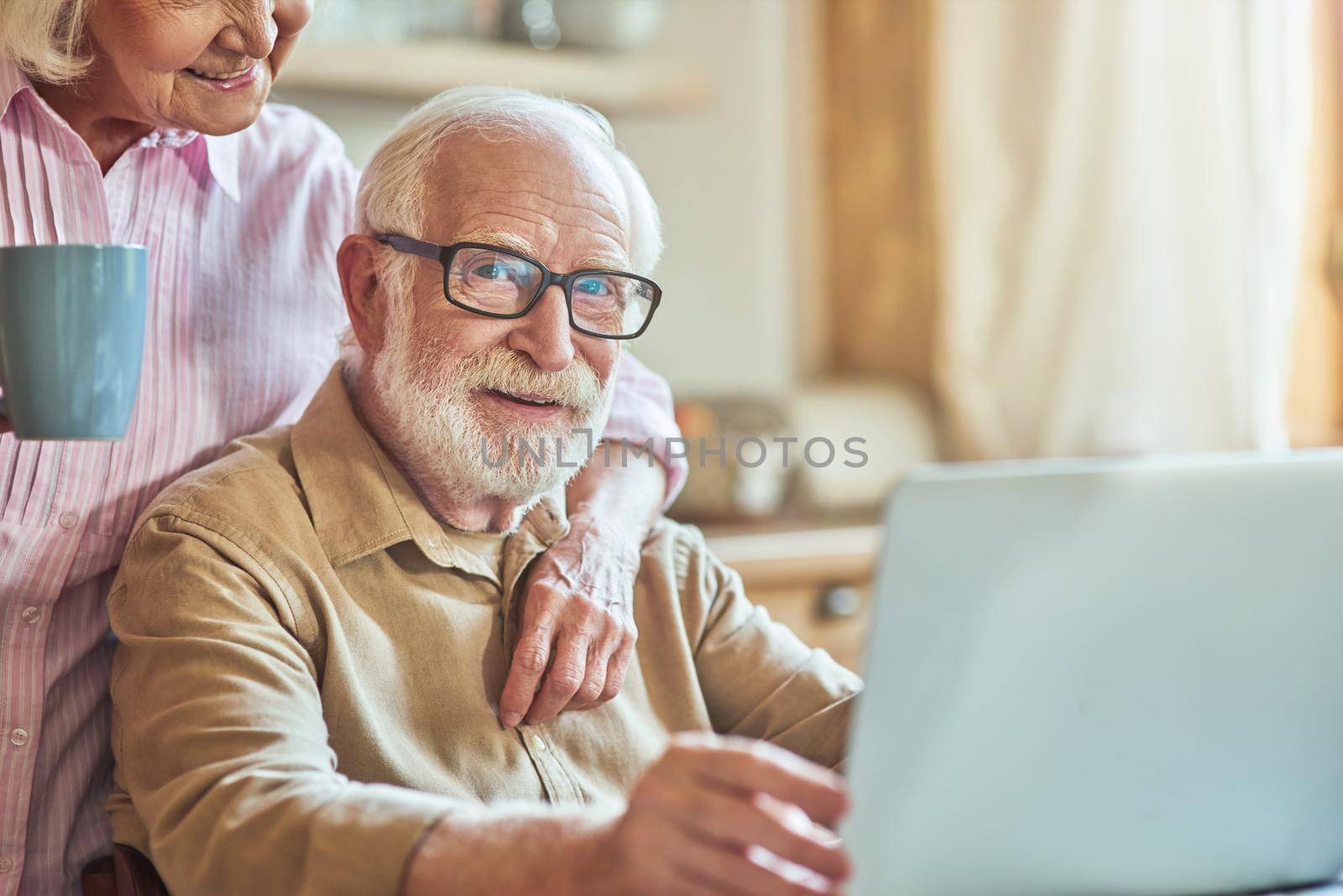 Happy elderly man in glasses working on laptop with her wife with cup standing near him at the kitchen. Domestic lifestyle concept