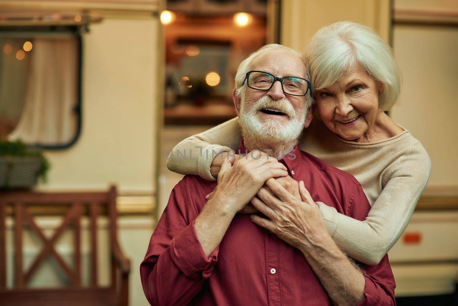 Happy senior woman hugging her husband while standing outdoors with their motorhome on the background. Travel concept