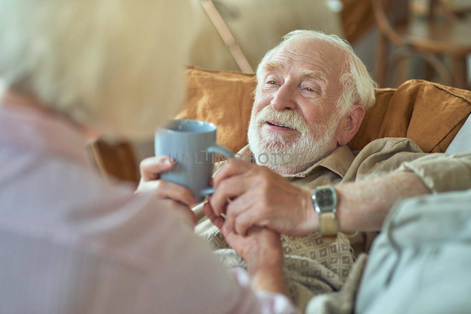 Smiling senior man sick at home and holding a cup of hot drink from his wife. Care and health concept