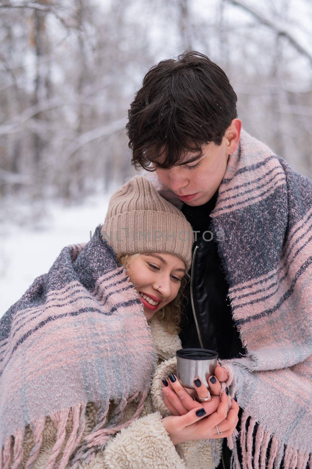 A young couple walks in the park in winter. The guy and the girl are drinking a warming drink outdoors