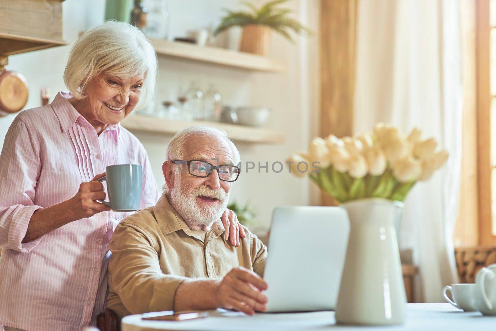 Smiling senior couple enjoying morning coffee while using laptop and watching video together. Domestic lifestyle concept