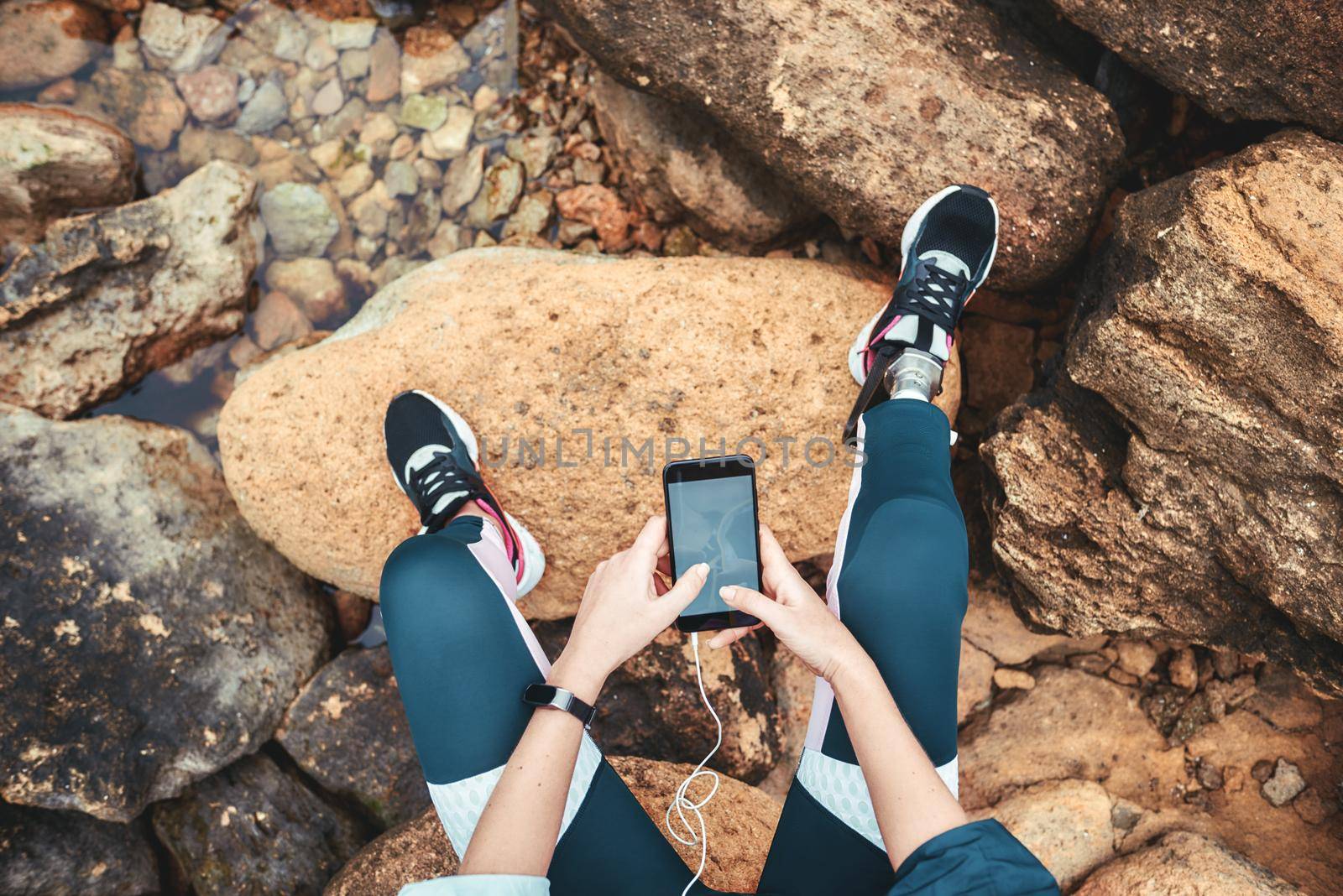 Top view of disabled woman in sport wear with leg prosthesis sitting on the boulders and holding smart phone by friendsstock