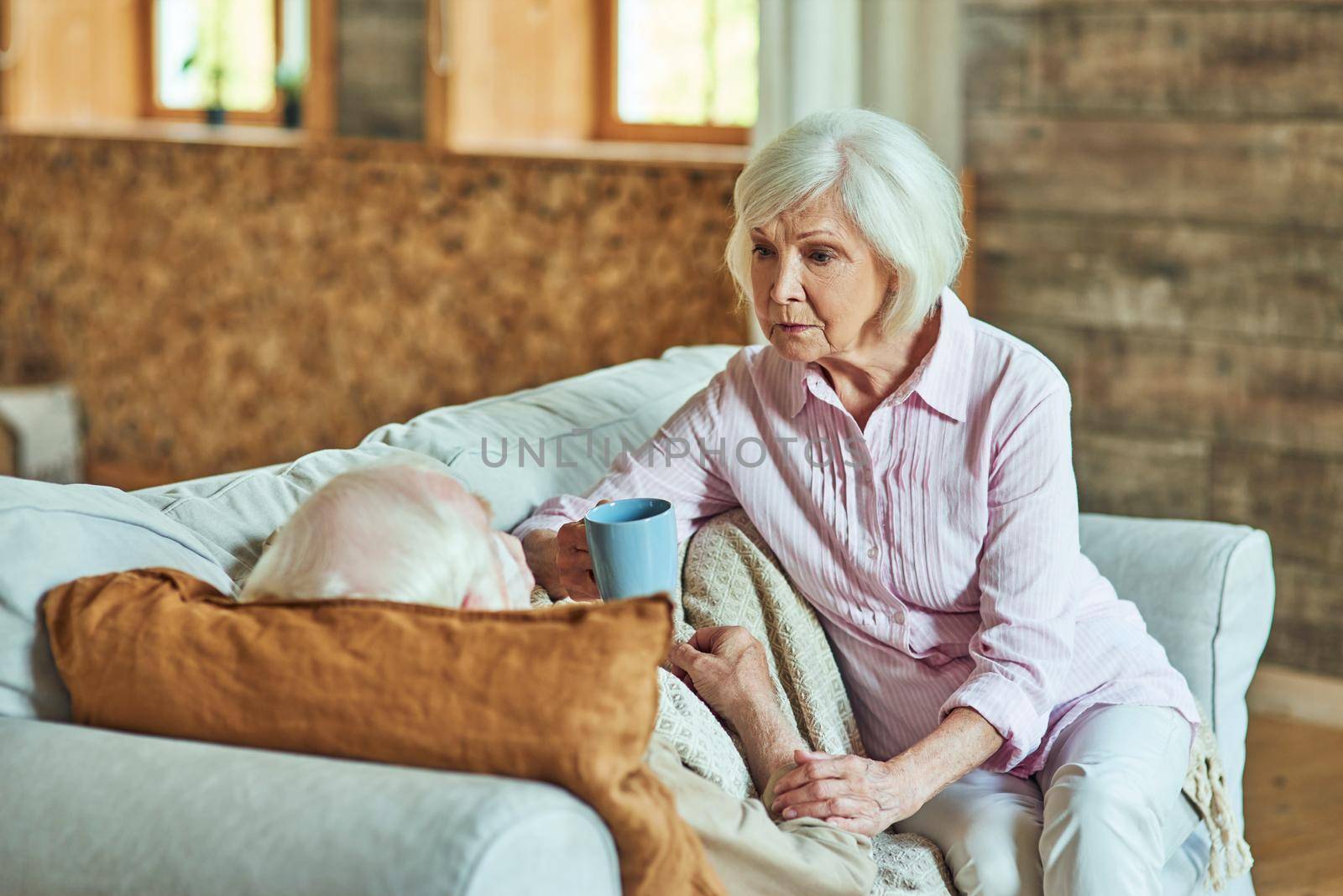 Elderly wife taking care of her husband while holding cup of tea and looking at him. Care and health concept