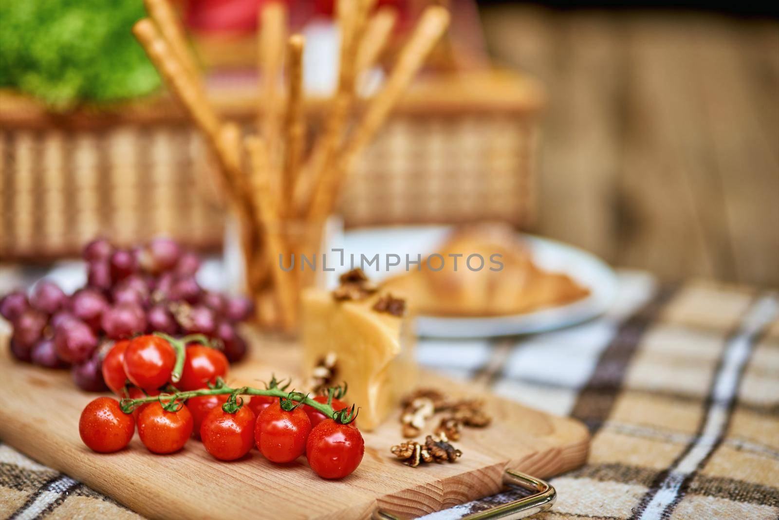 Tomatoes, grapes and cheese on a cutting wooden board on checkered plaid with a wicker basket on the background. Rest and vacation lifestyle concept
