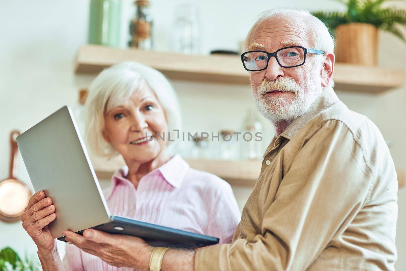 Smiling senior couple standing at the kitchen while using laptop and looking at camera at their home. Lifestyle concept