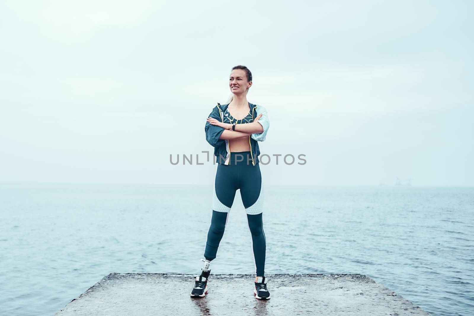 Feeling happy. Positive and motivated disabled woman in sports clothing with prosthetic leg standing with crossed arms on the beach and smiling. by friendsstock