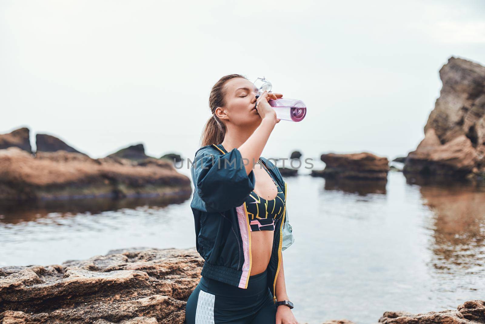 Water is life. Sporty young woman in sportswear drinking water while sitting on the stone at the beach. Motivation. Sport concept. Taking a break