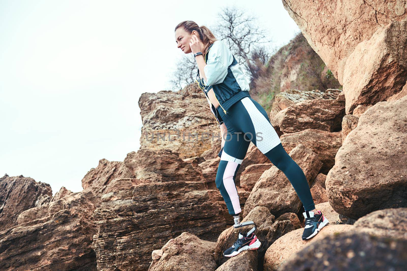 Positive disabled woman in sport wear with leg prosthesis holding phone in her hand and listening music while standing on the boulders. Digital concept. Workout. Disabled Sportsman. Healthy lifestyle.