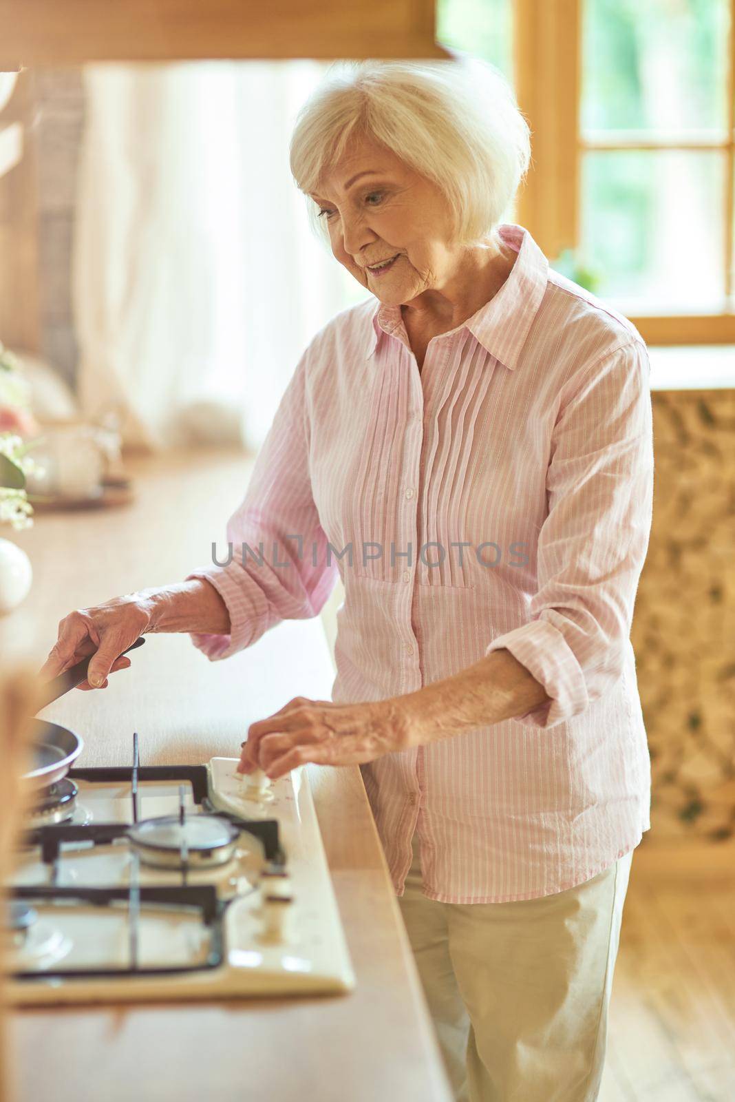Smiling senior woman turning on the stove and setting the frying pan to make breakfast at home. Domestic lifestyle concept
