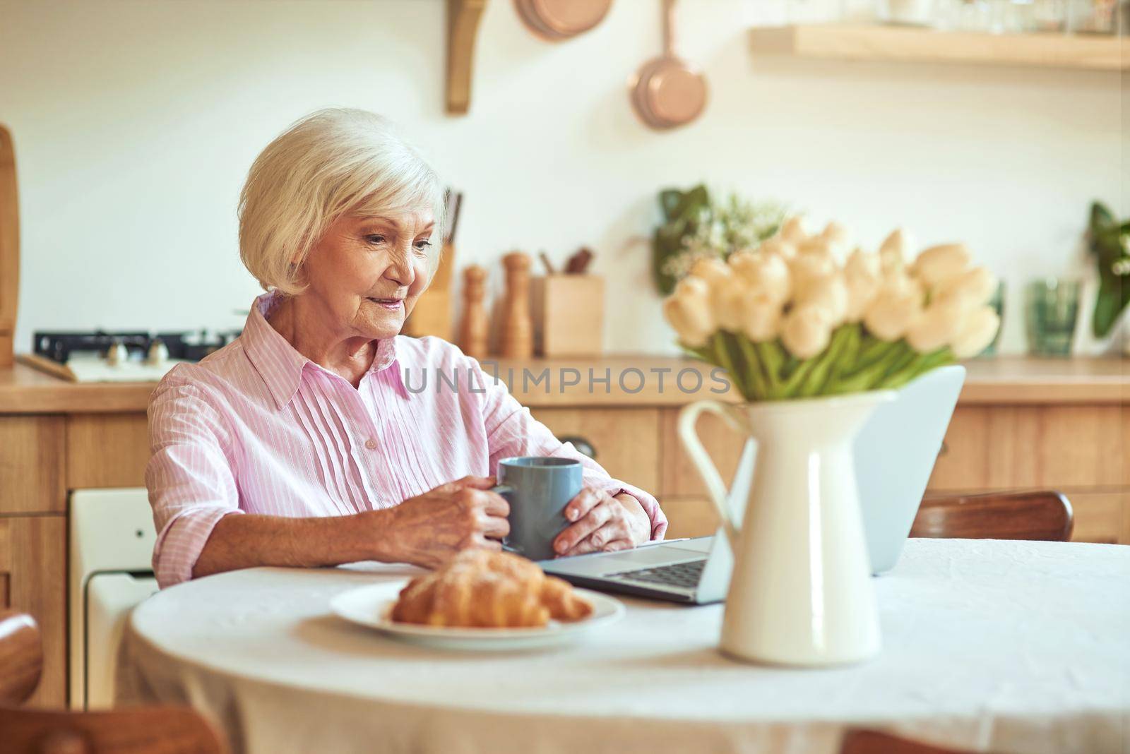 Happy beautiful senior lady holding cup of coffee and looking at laptop screen at the kitchen. Domestic lifestyle concept