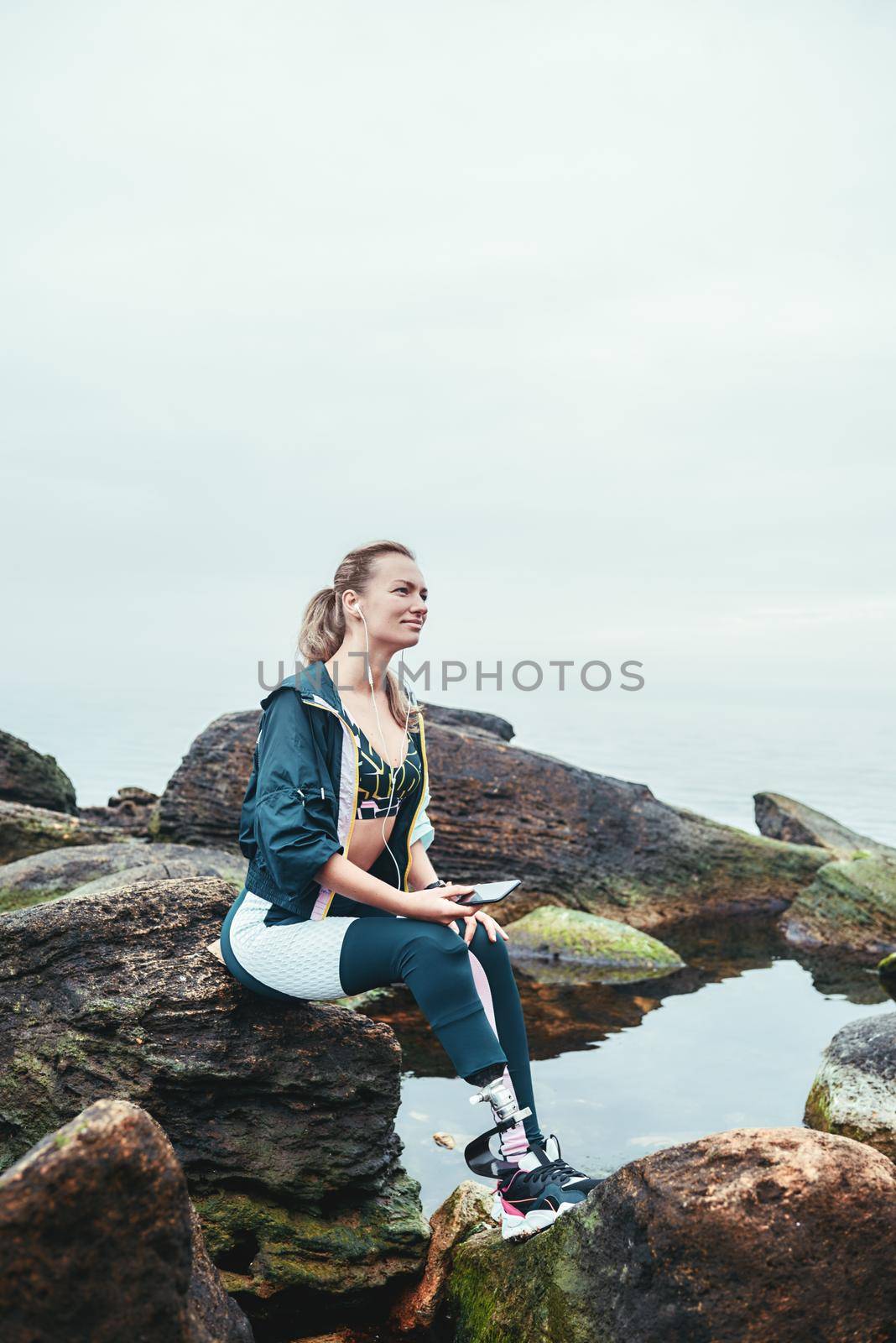 Enjoying nature and music. Happy disabled athlete woman in sportswear with prosthetic leg sitting on the stone and listening music. Sport concept. Music concept. Disabled Sportsman.