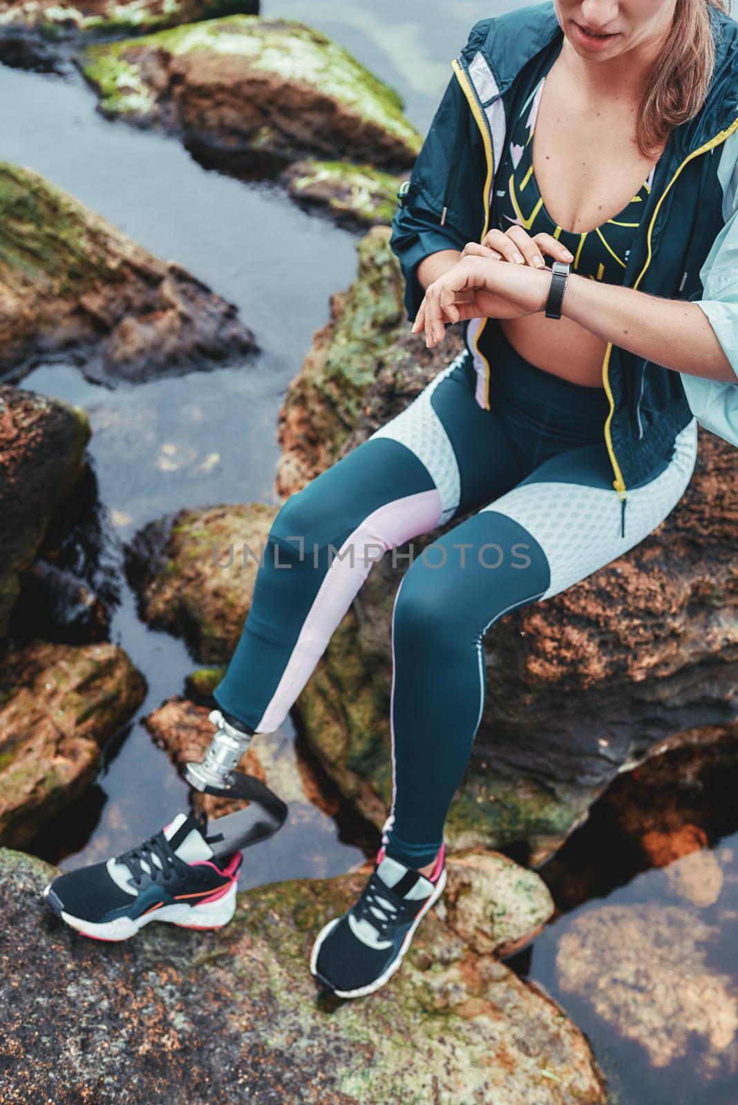 Checking results. Cropped photo of disabled athlete woman in sportswear with prosthetic leg checking pedometer while sitting on the stone by friendsstock