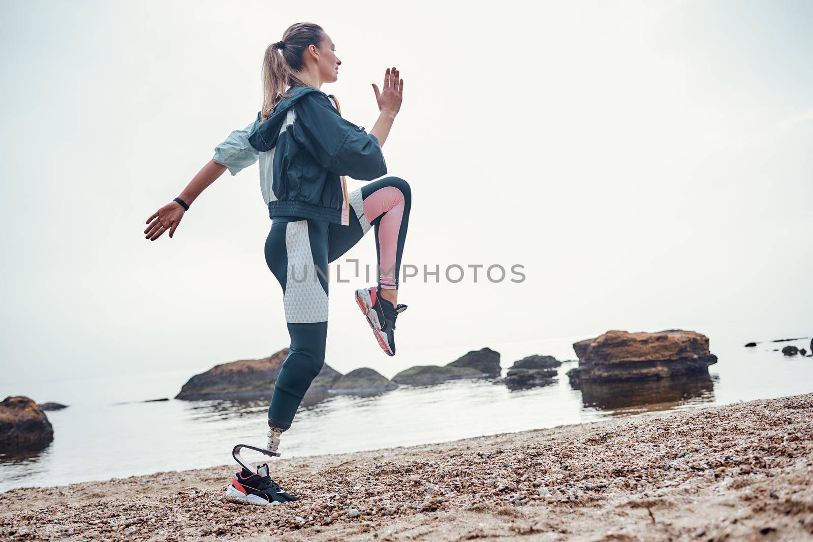 Exercising every morning. Side view of young sporty disabled woman with prosthetic leg is doing sport exercises on the beach by friendsstock
