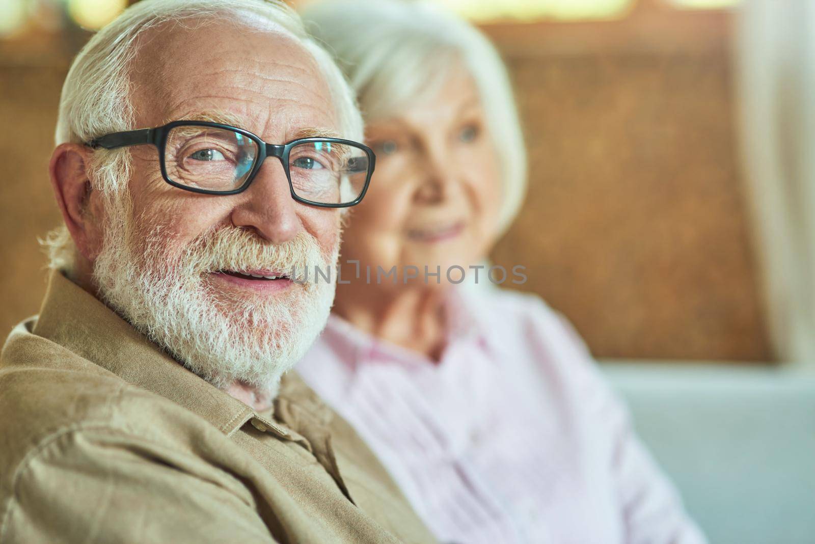 Smiling senior man wearing glasses looking at camera with her wife sitting on the background at home. Family and relationships concept