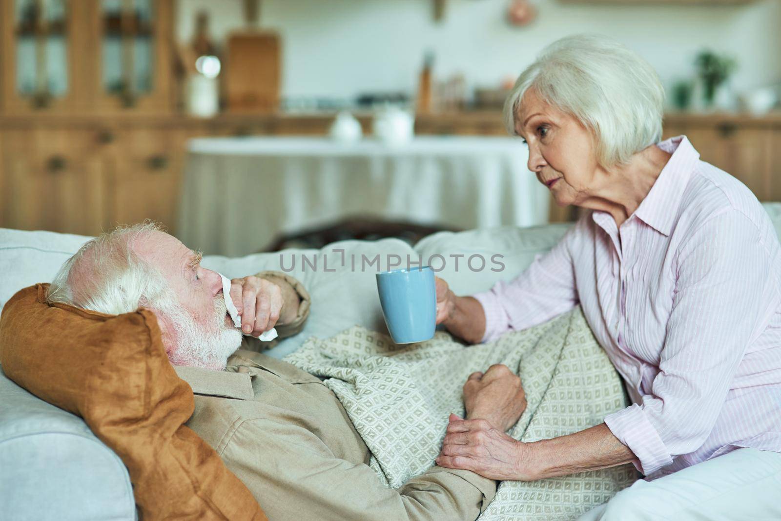 Senior woman taking care of her spouse while holding cup of hot drink and sitting next to him. Care and health concept