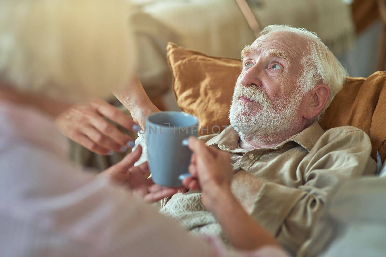 Elderly gray-haired man sick at home while his wife caring for him and holding hot drink for him. Care and health concept