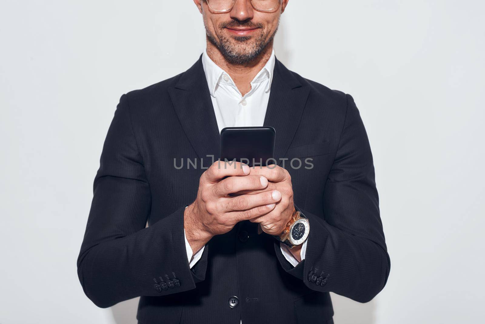 Great news. Portrait of handsome and cheerful bearded man in classic wear and eyeglasses holding smart phone and looking at it while standing against grey background. Digital concept. Business concept