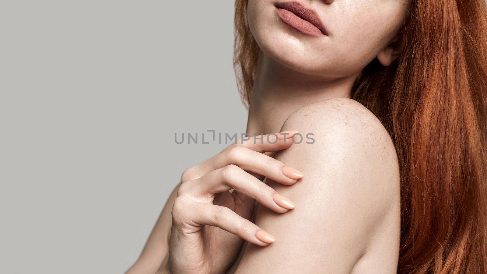 Studio shot of beautiful young redhead woman touching her soft skin while standing against grey background