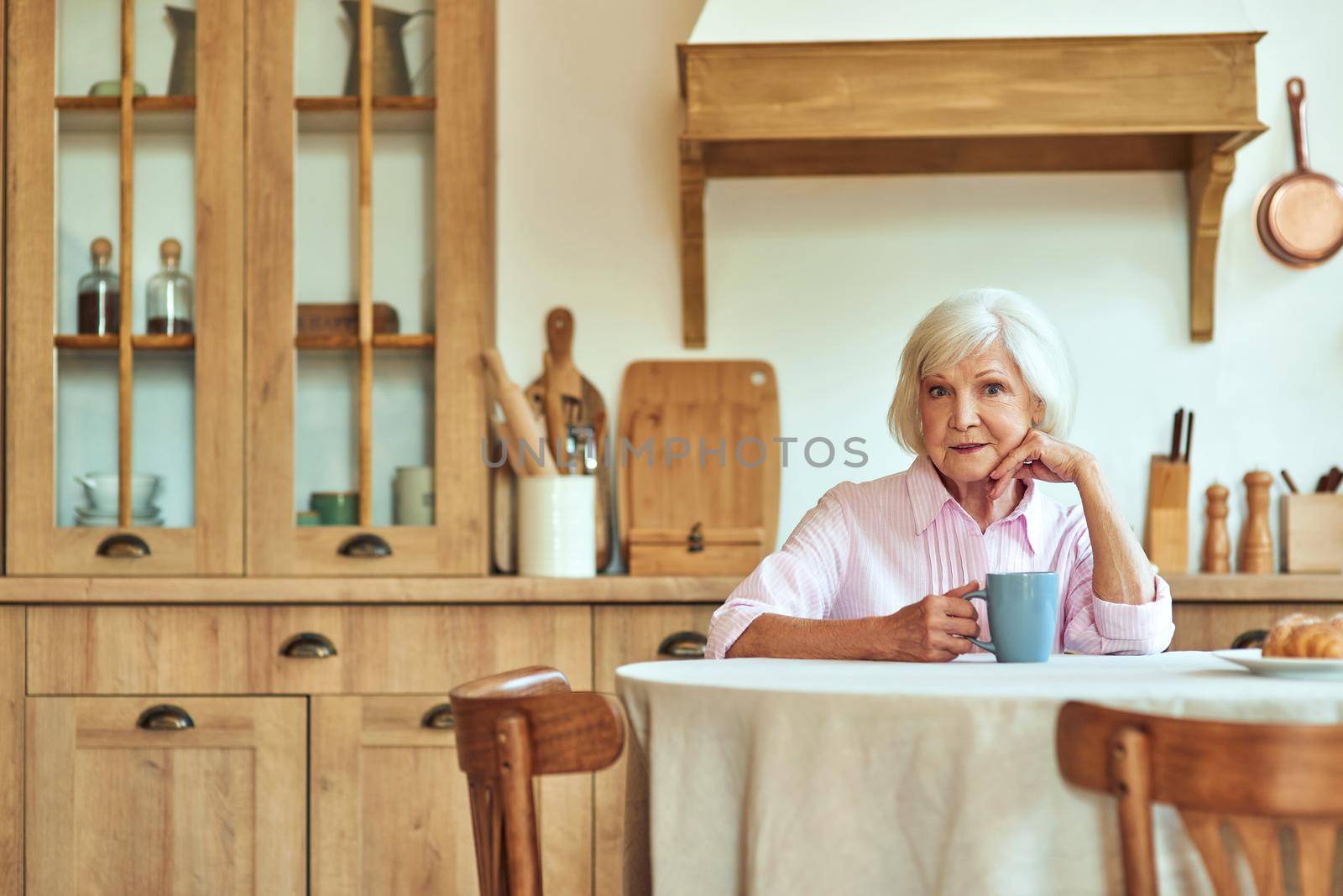 Cheerful elderly woman with cup of hot drink sitting at the kitchen by friendsstock