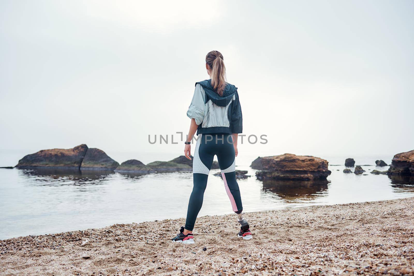 I adore living by the sea. Back view of confident disabled sporty woman in sportswear with prosthetic leg is standing on the beach by friendsstock