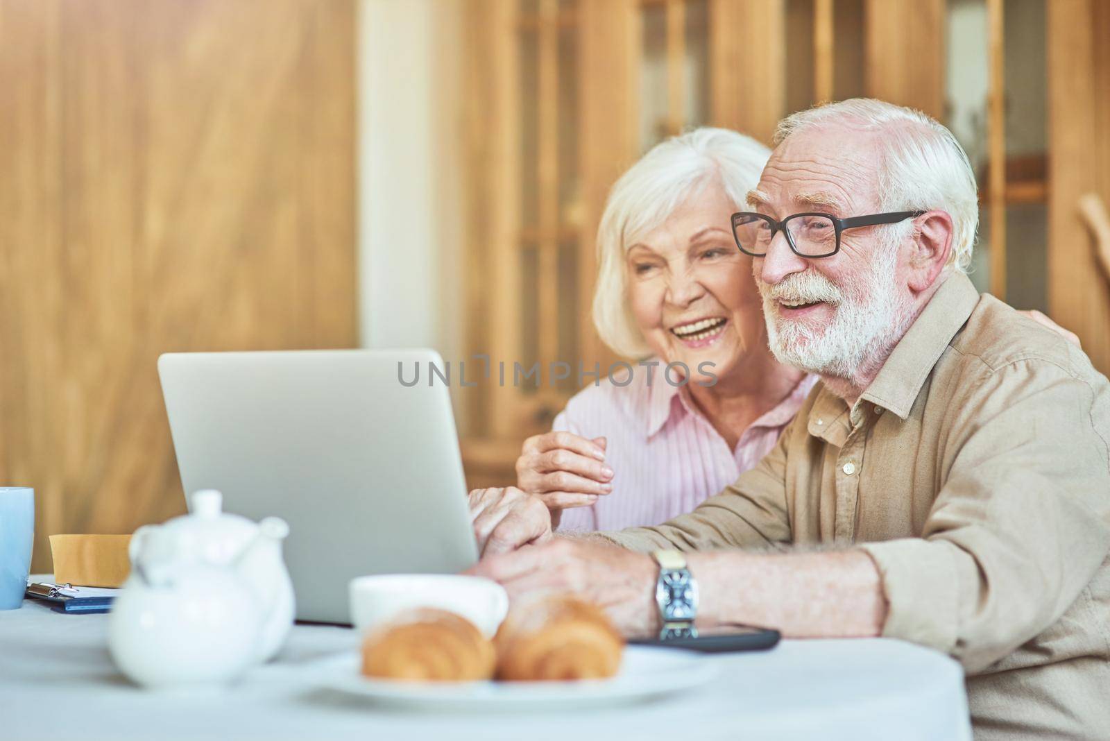 Happy senior man and woman hugging while sitting at the table and looking at laptop screen. Domestic lifestyle concept