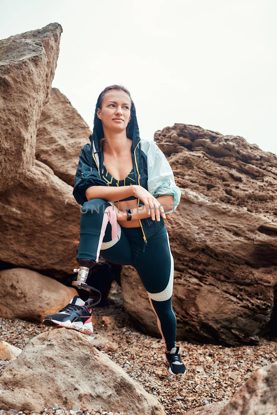 After workout. Vertical photo of disabled athlete woman in sportswear with prosthetic leg standing on the beach and looking at camera. by friendsstock