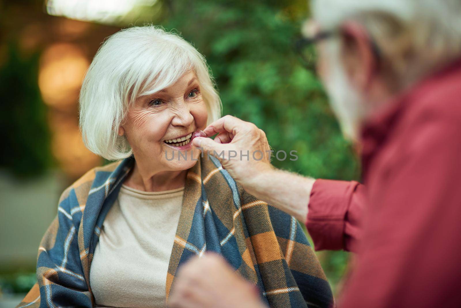 Cropped photo of elderly man treating his wife to grapes while sitting together outdoors. Relationship and family concept