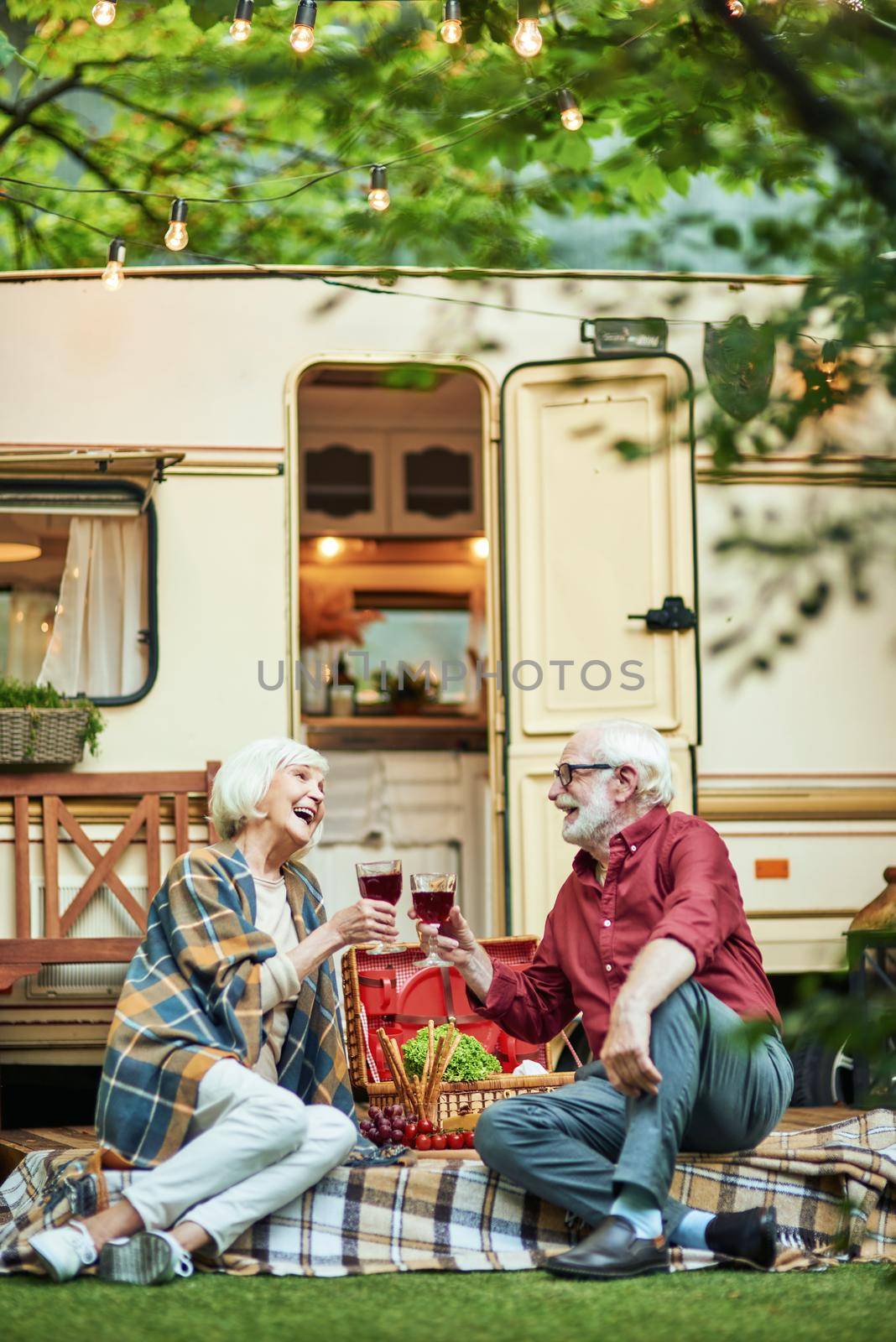 Full length photo of senior couple flirting and laughing together while holding wine near their camper van. Travel concept