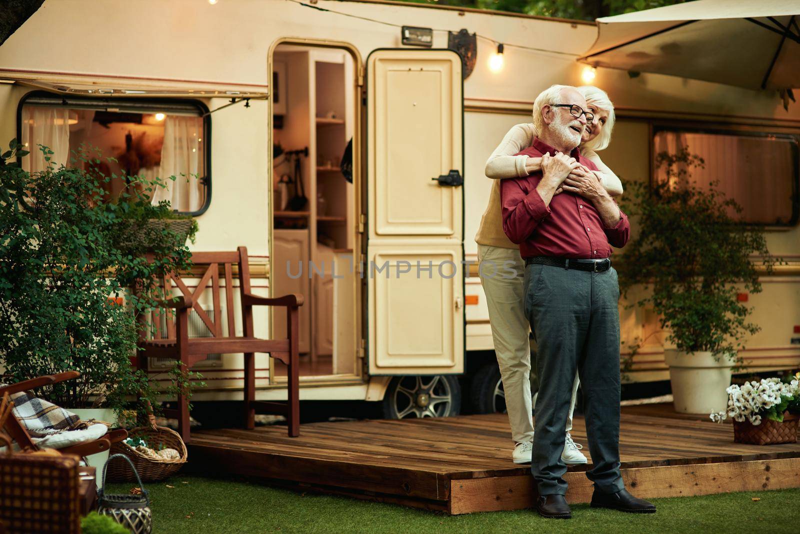 Full length photo of smiling elderly married couple hugging while standing on the van porch. Travel concept