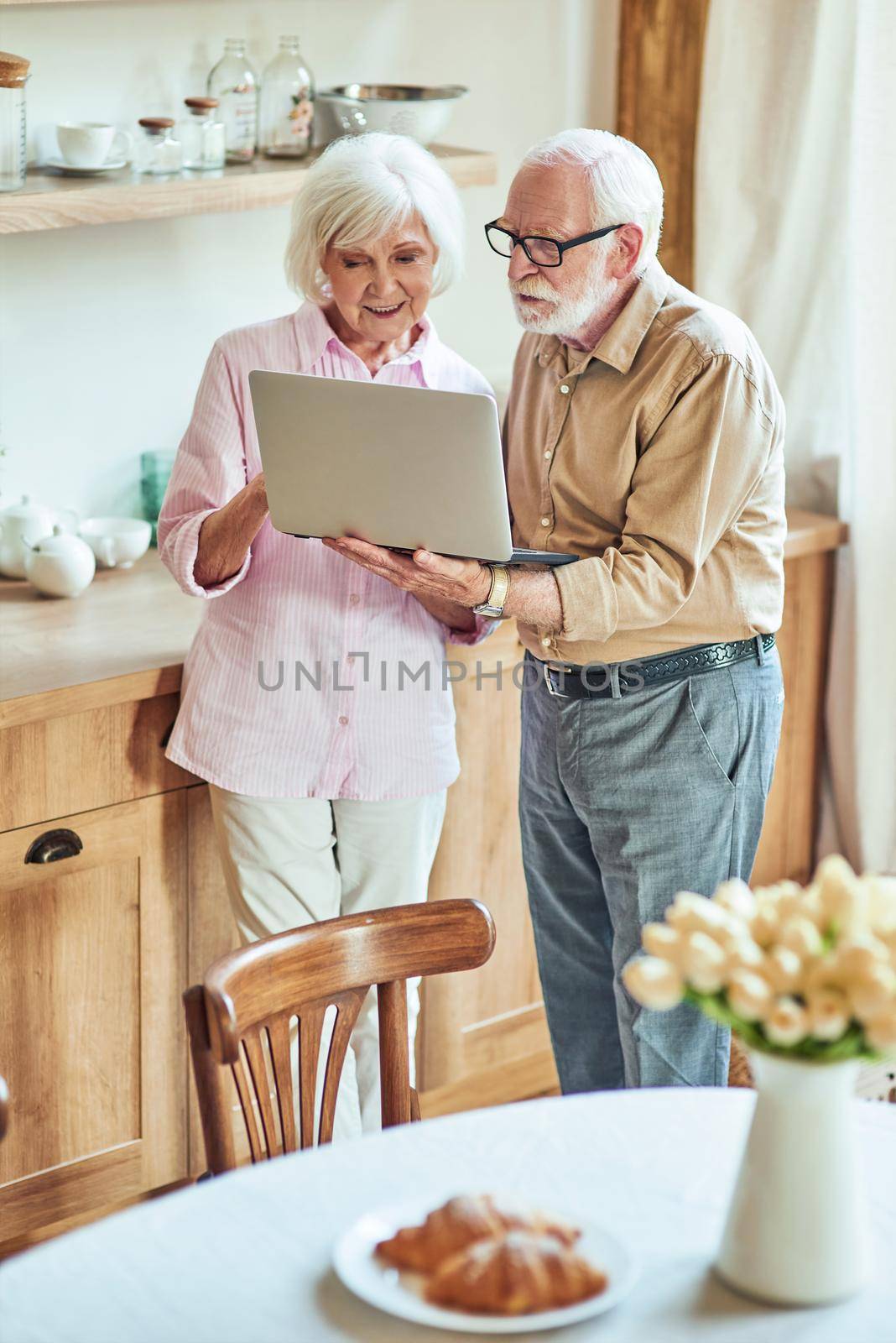 Happy senior couple browsing the internet together at the kitchen by friendsstock