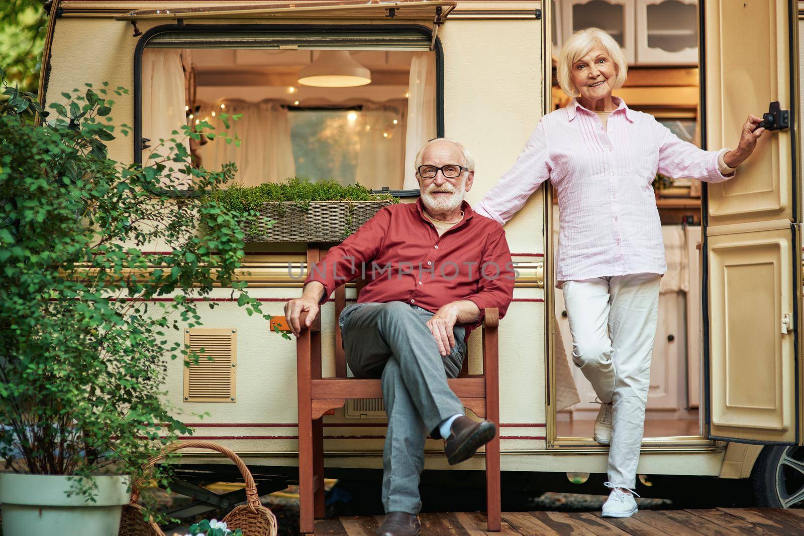Smiling senior man sitting on wooden chair on the porch of his motorhome while his wife standing next to him. Travel concept