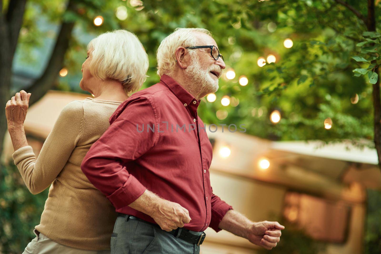 Smiling gray-haired man with glasses dancing with his wife back to back by friendsstock