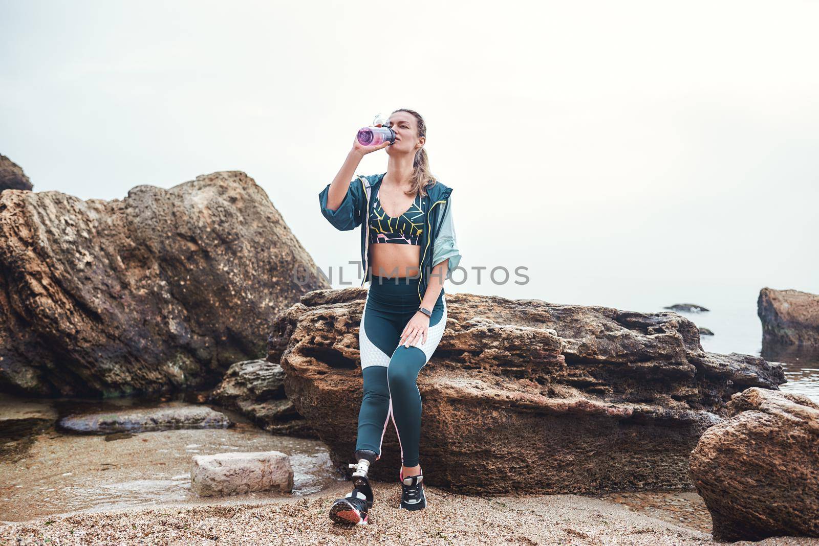 Taking a break. Tired disabled athlete woman with prosthetic leg drinking water while sitting on the stone at the beach by friendsstock
