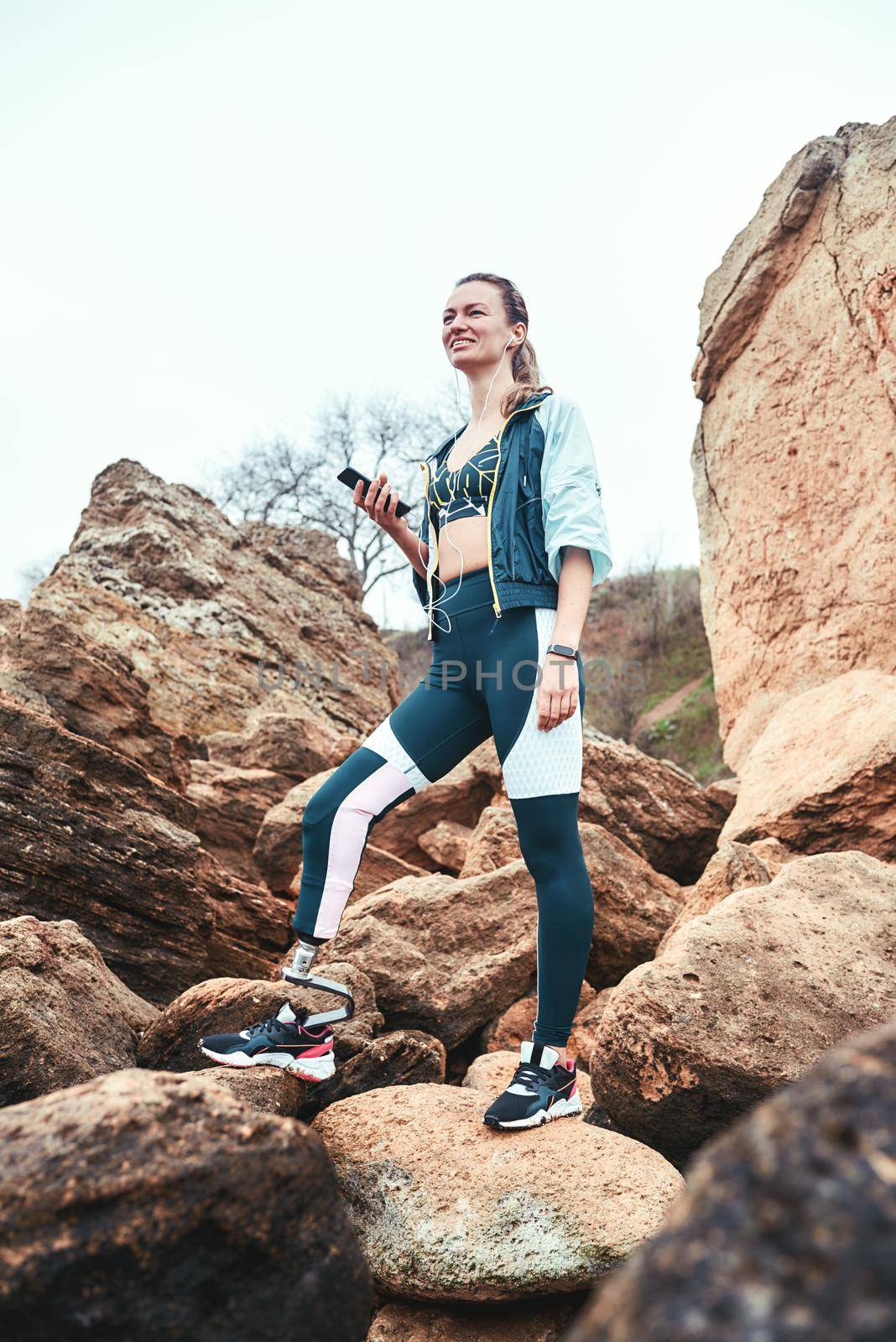 Resting after workout. Disabled woman in sport wear with leg prosthesis holding phone in her hand and listening music while standing on the boulders. Digital concept. Workout. Disabled Sportsman. Healthy lifestyle.