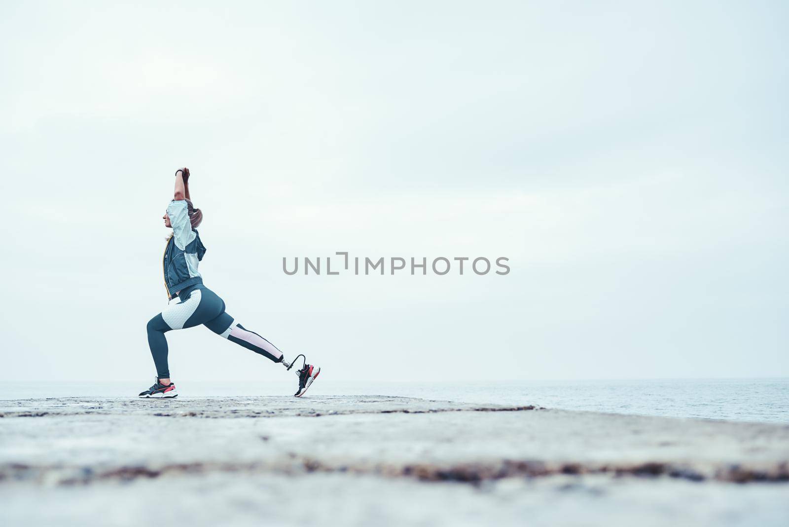 My perfect morning. Sporty disabled athlete woman in sportswear with prosthetic leg standing in yoga pose on the stone in front of the sea. by friendsstock