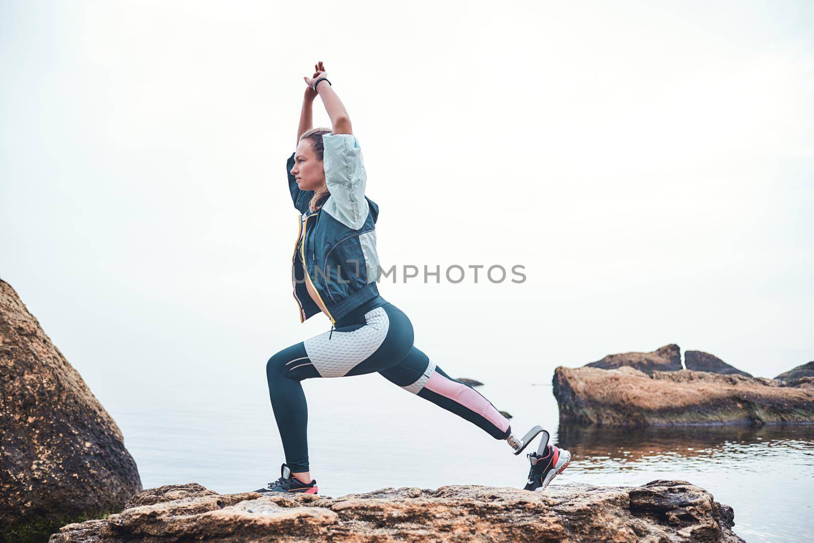 Life balance. Calm and sporty disabled athlete woman in sportswear with prosthetic leg standing in yoga pose on the stone in front of the sea. Sport concept. Disabled Sportsman. Healthy lifestyle