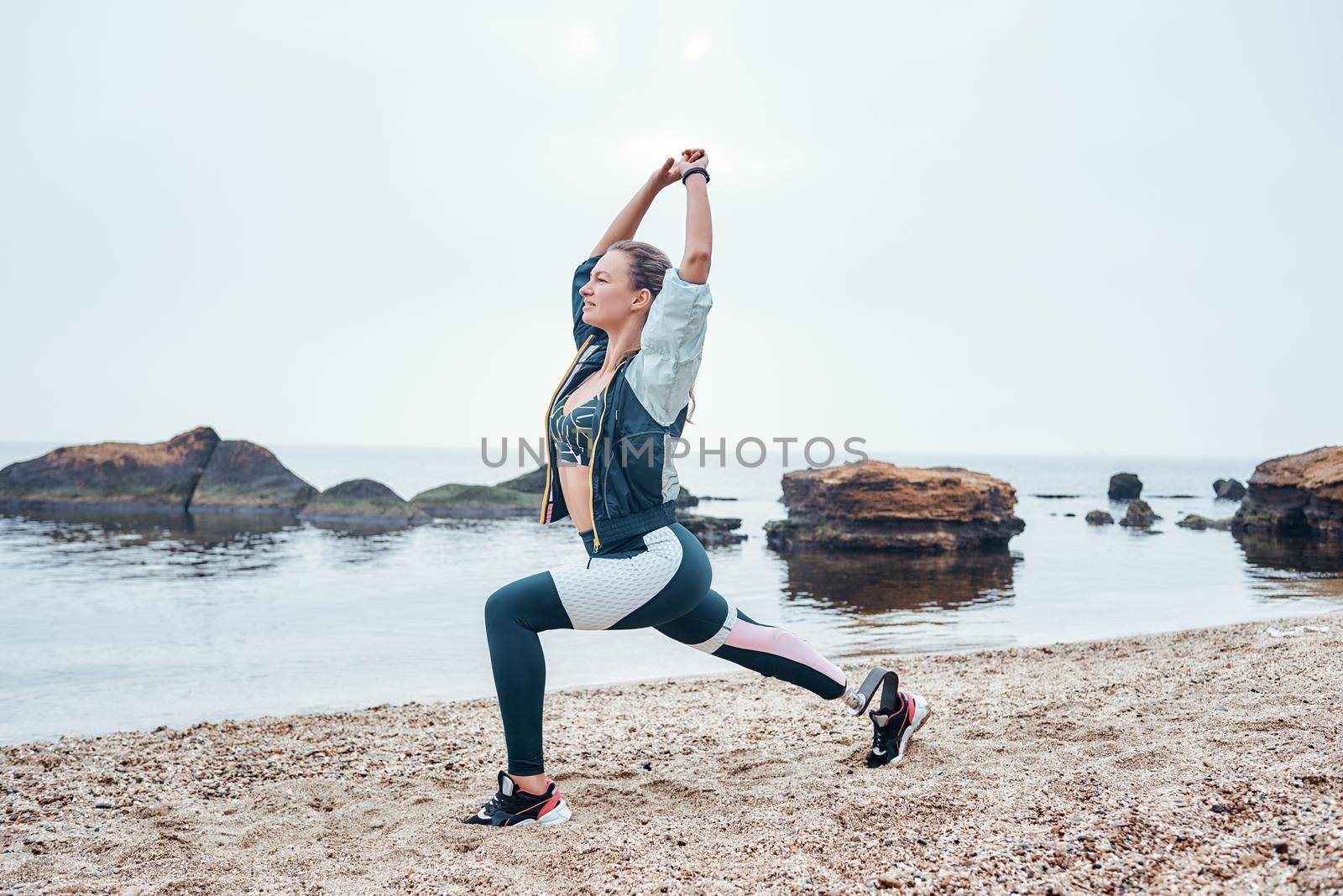 Exercises with sunrise. Side view of beautiful disabled athlete woman in sportswear with prosthetic leg standing in yoga pose at the beach. by friendsstock