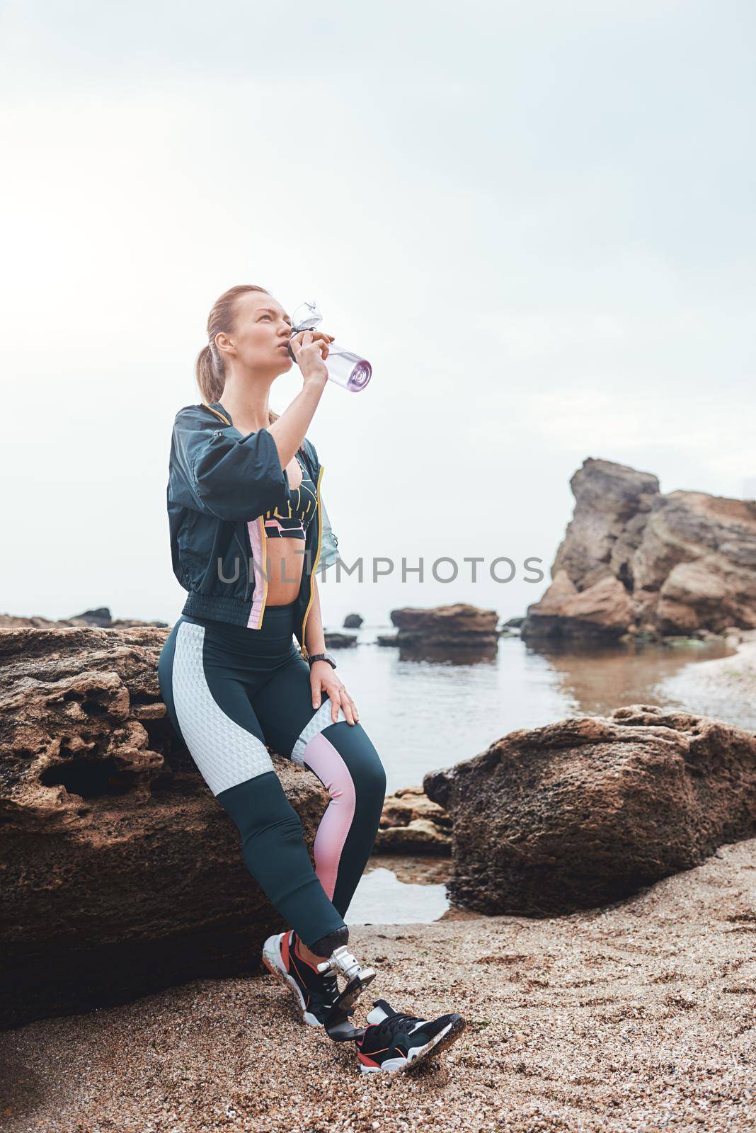 I need some water. Sporty and healthy disabled athlete woman with prosthetic leg drinking water while sitting on the stone at the beach. Disabled Sportsman. Motivation. Sport concept. Taking a break