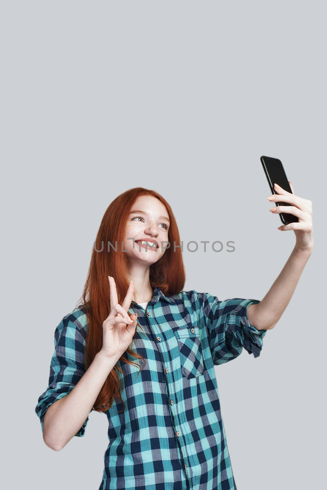 Peace Cute young redhead woman in casual wear making selfie and gesturing while standing against grey background. Positive emotions. Digital concept. Photography