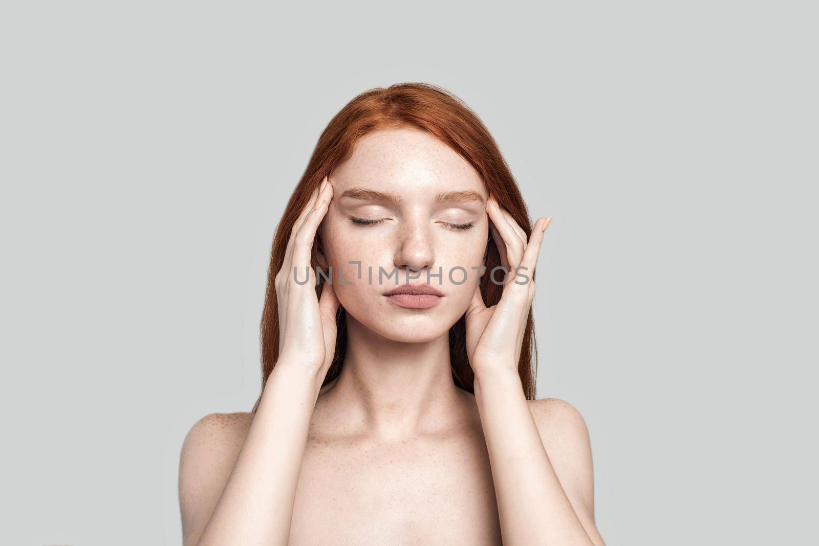 Studio shot of beautiful redhead woman keeping eyes closed and touching head with hands while standing against grey background