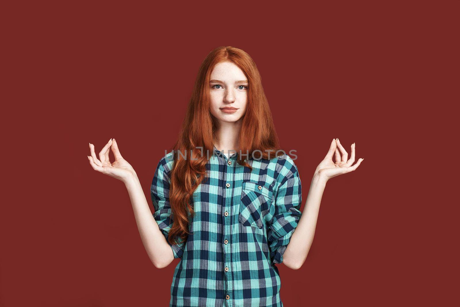 Photo of relaxed beautiful female in checkered shirt, making yoga gesture, isolated over red studio background. People, meditation, wellness, relaxation and lifestyle concept
