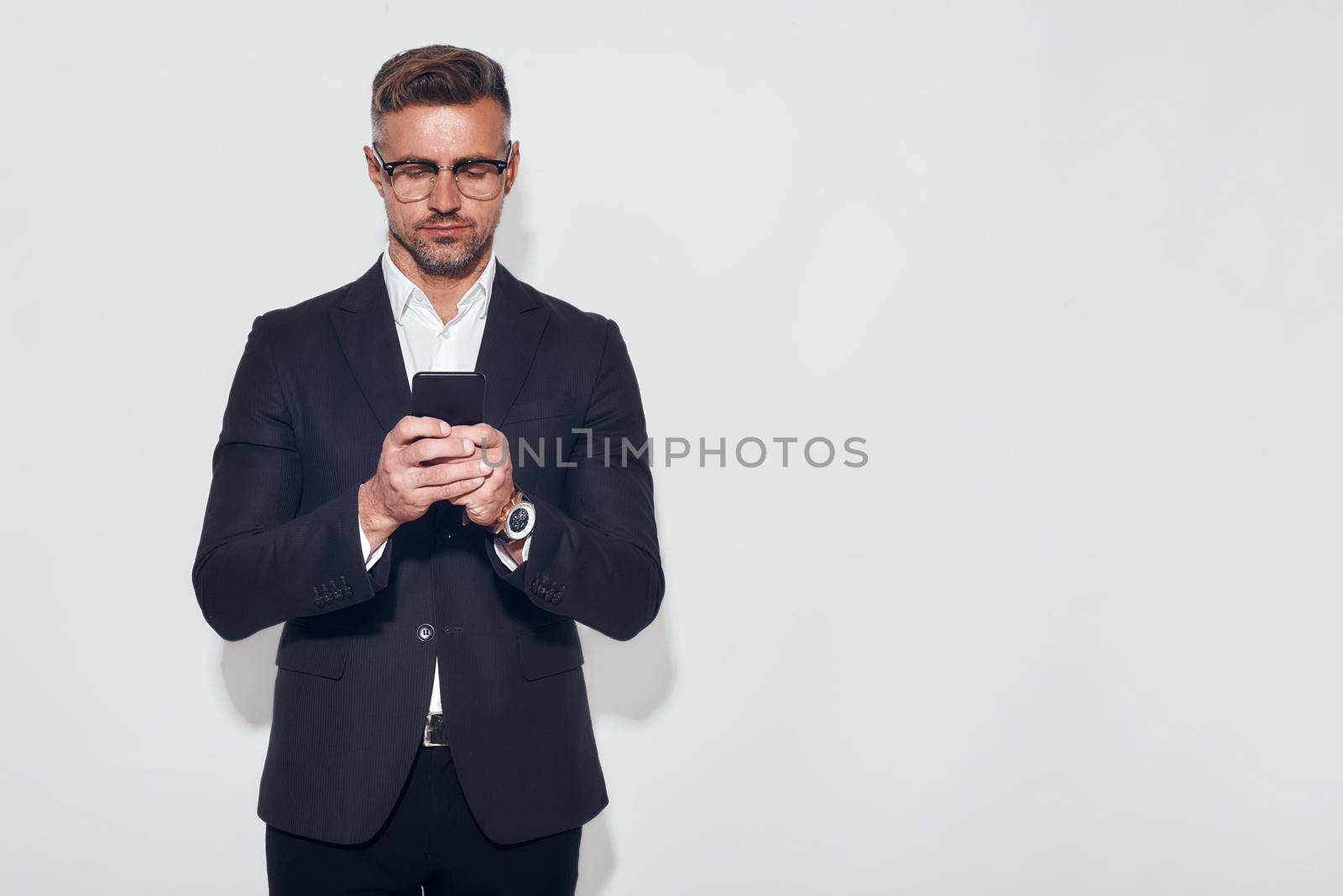 Always in touch. Portrait of handsome bearded man in classic wear and eyeglasses holding smart phone and looking at it while standing against grey background. Digital concept. Business concept