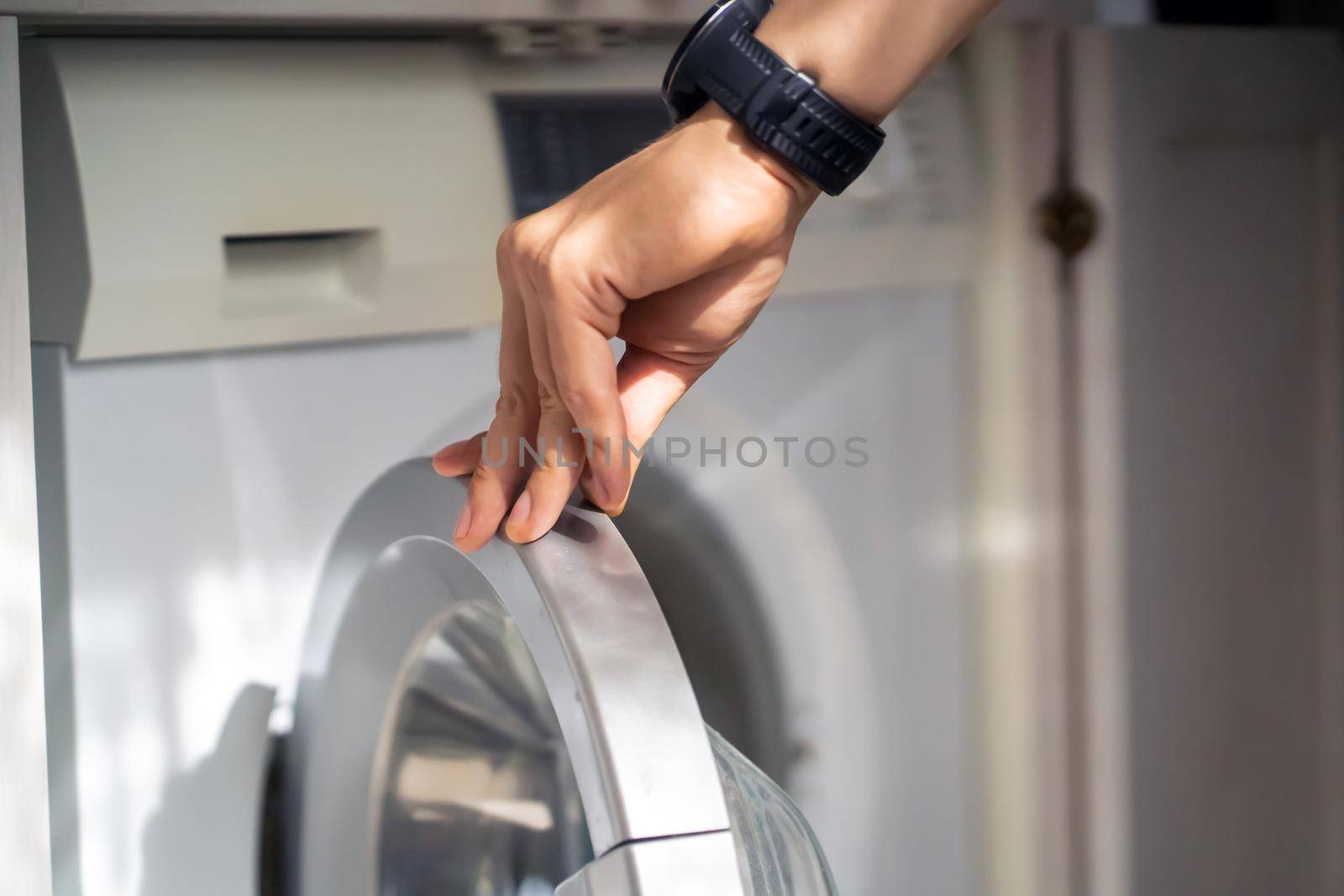A male hand opens the door of a new washing machine in his modern, bright bathroom, close-up. The man washes clothes in the laundry.