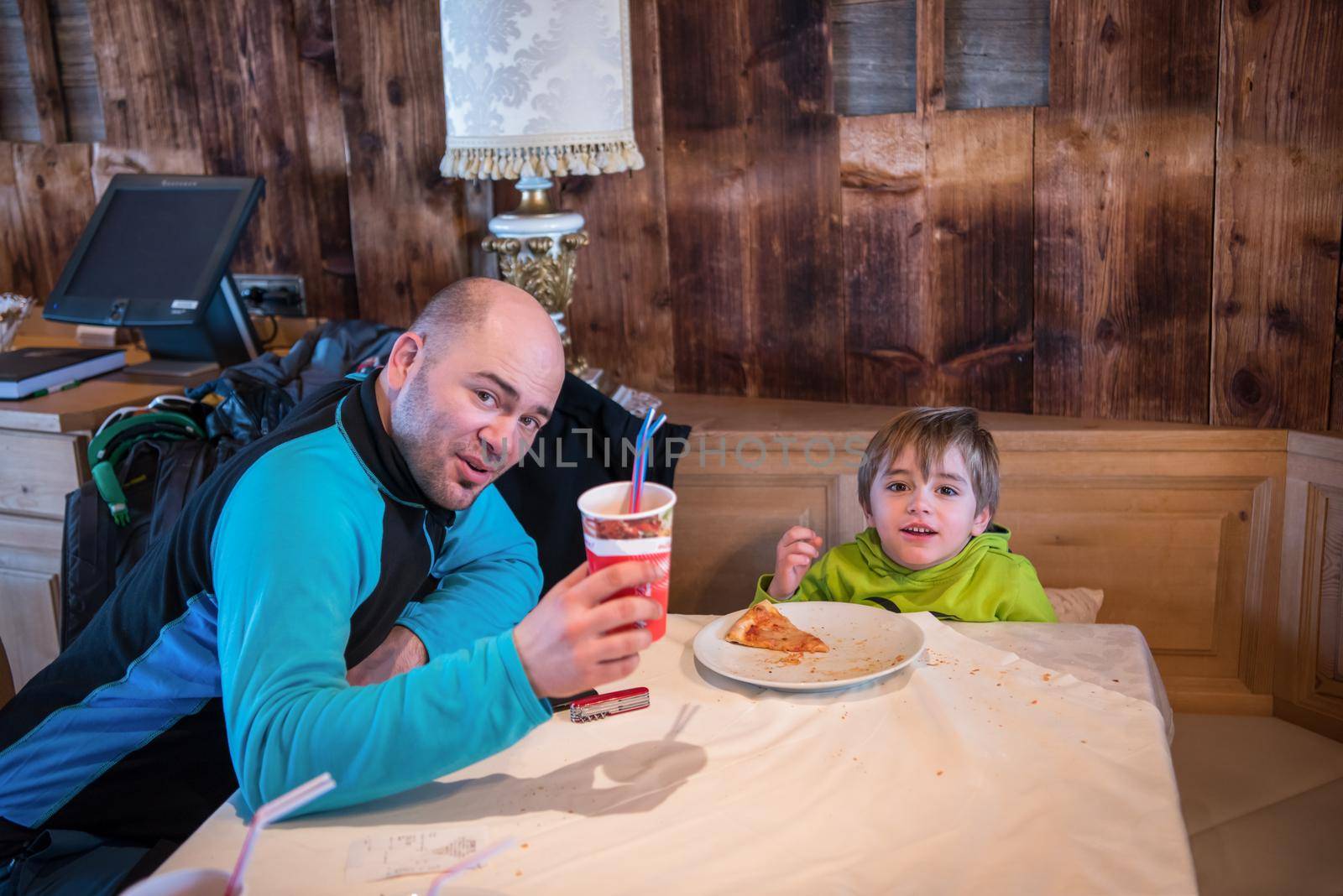 young happy father and his little son eating a pizza after a hard snowboarding day during winter vacation at beautiful ski resort