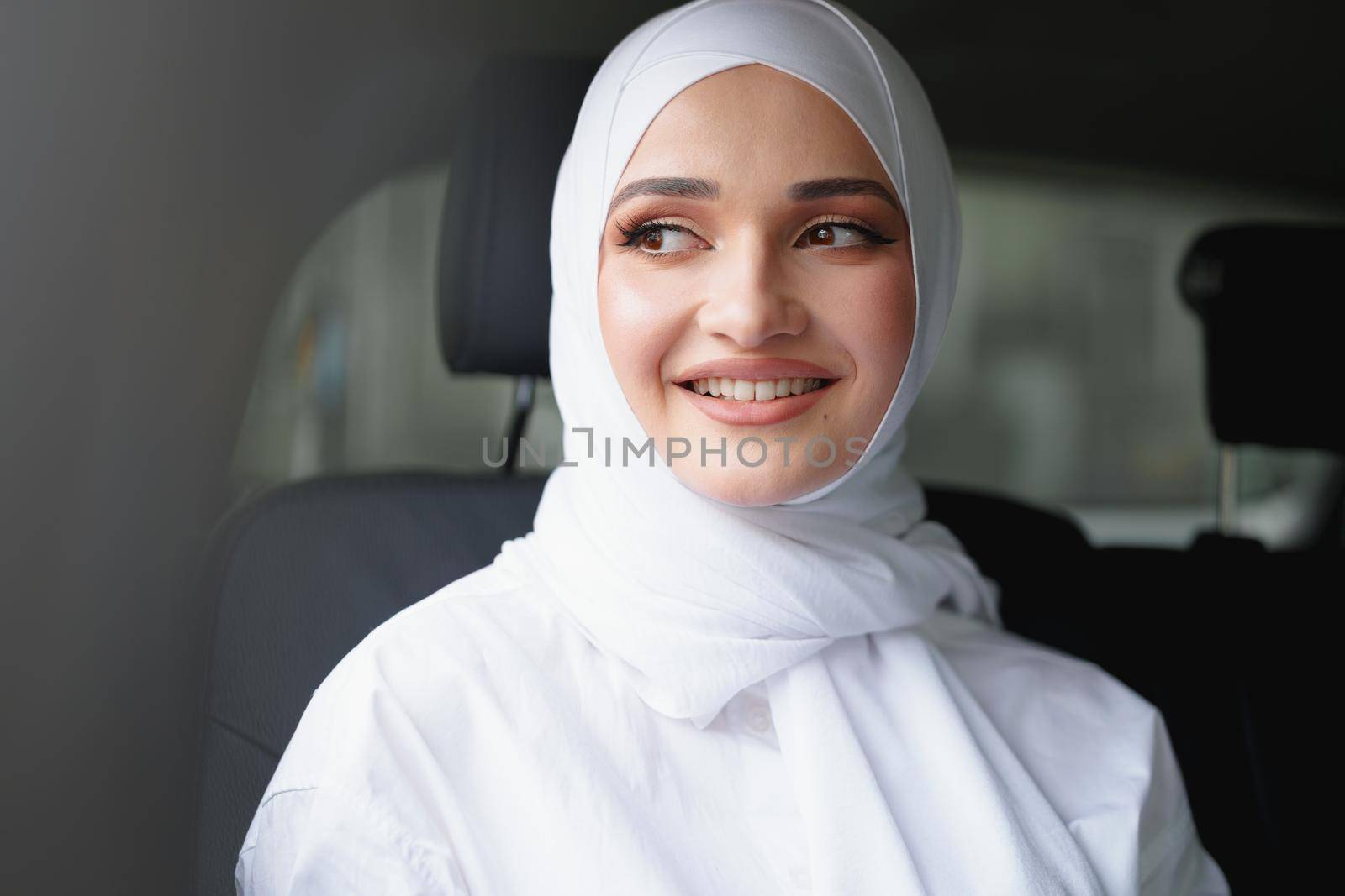 Beautiful muslim woman in white hijab sitting on the back seat of a luxury car, portrait