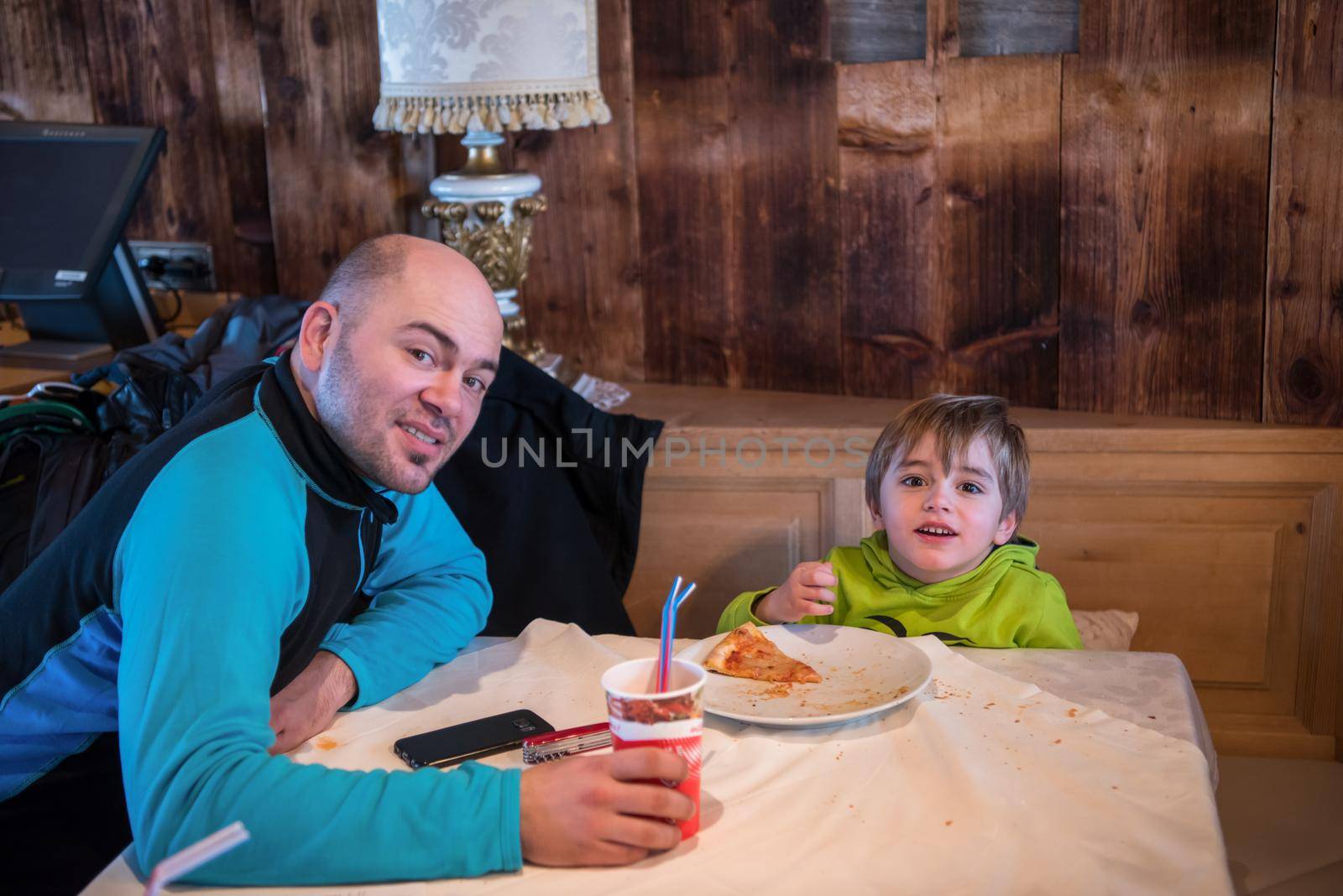 young happy father and his little son eating a pizza after a hard snowboarding day during winter vacation at beautiful ski resort