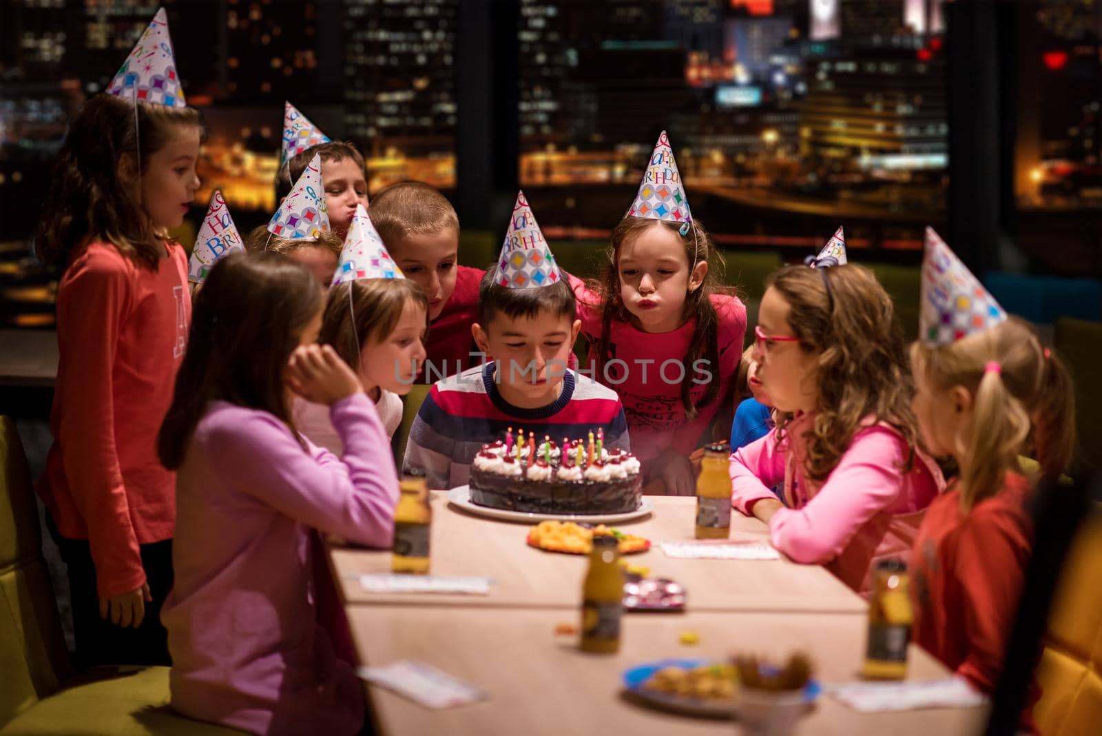 young happy boy and group of his friends having birthday party with a night city through the windows in the background