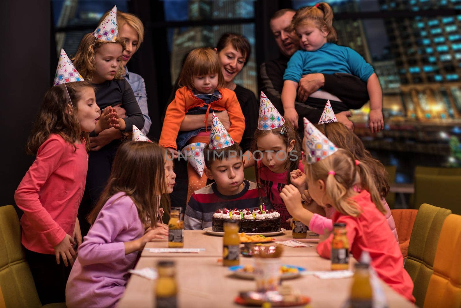 young happy boy and group of his friends having birthday party with a night city through the windows in the background