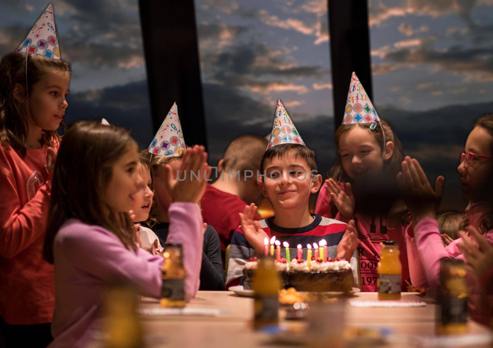 young happy boy and group of his friends having birthday party with a night sky through the windows in the background