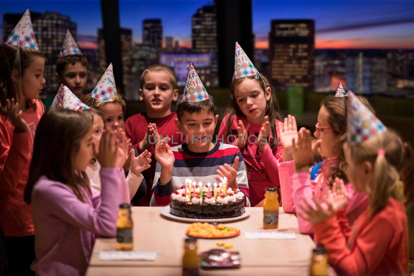 young happy boy and group of his friends having birthday party with a night city through the windows in the background